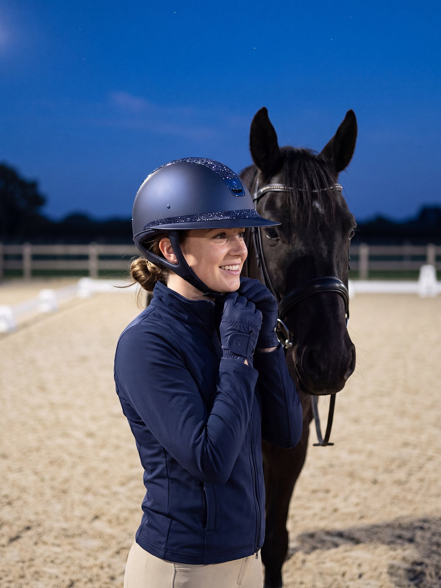Rider wearing a navy Samshield Miss Shield 2.0 Cosmic Night helmet putting on gloves at a floodlit arena at dusk